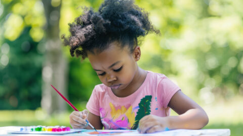 Young child with dark skin painting colorfully on large paper, smiling with joy